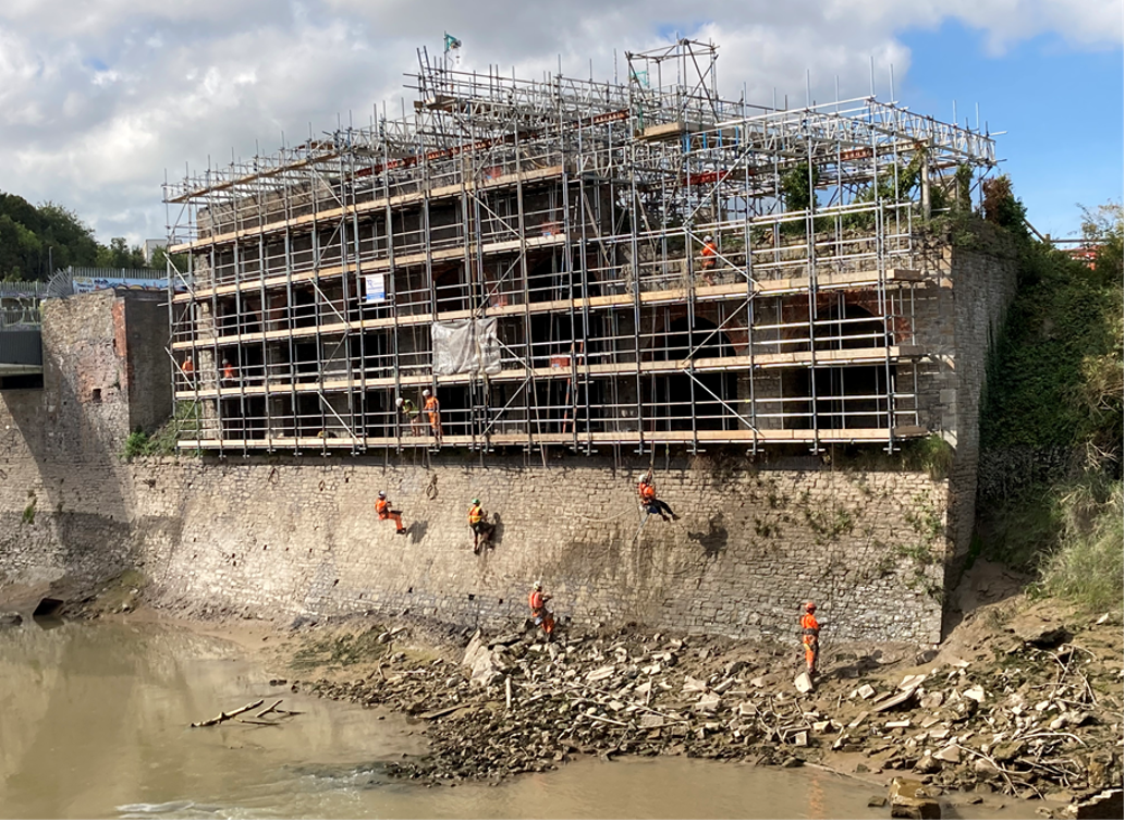 River wall at Temple Island covered in scaffolding. People in high vis clothing abseil down the wall while repairing it.